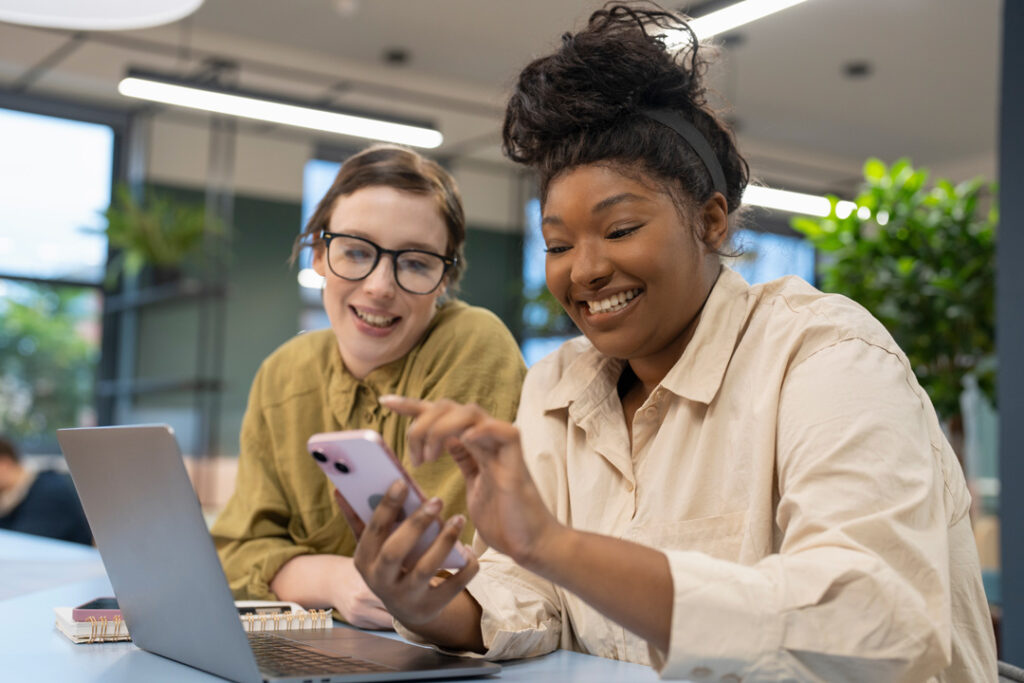 smiling businesswomen using phone and laptop in office