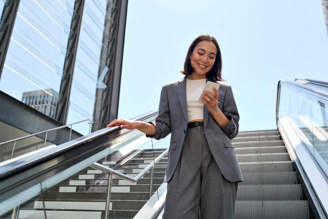 smiling young asian business woman wearing suit standing on urban escalator using applications on cell phone